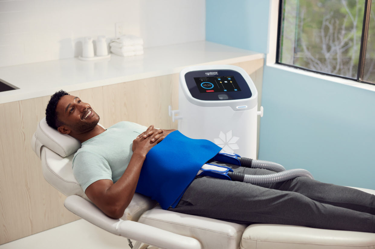 A man is lying on a treatment chair with CoolTone applicators attached to his abdominal area, while a CoolTone device is positioned beside him in a modern medical setting, highlighting the benefits of this non-invasive muscle-toning treatment.