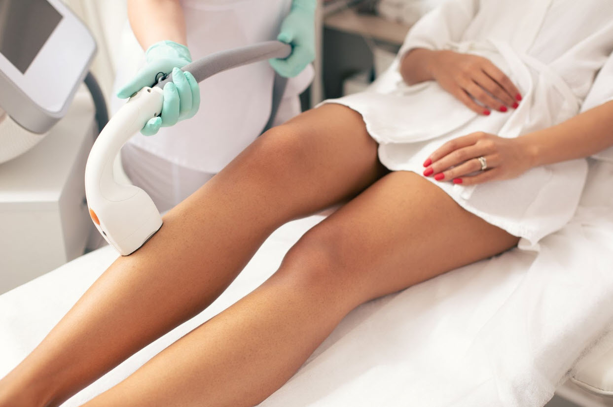 A woman in a white robe is lying on a treatment bed while a professional wearing gloves performs a laser hair removal procedure on her legs, emphasizing the importance of preparing for laser hair removal to achieve optimal and long-lasting results.